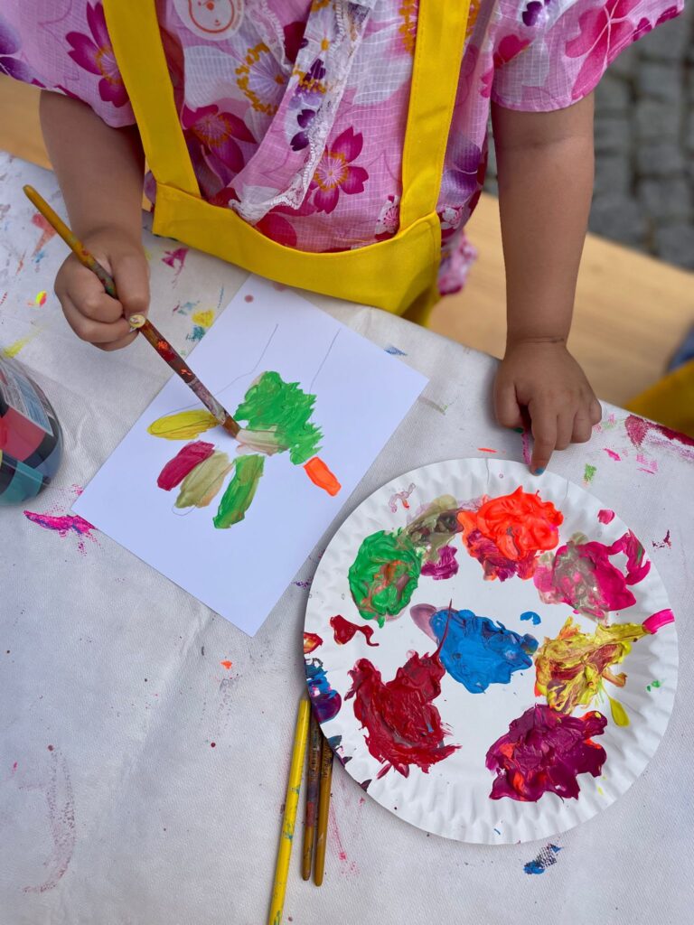 Ein Kind malt eine Hand auf ein Blatt Papier. Neben dem Kind liegt eine Palette mit Farben.