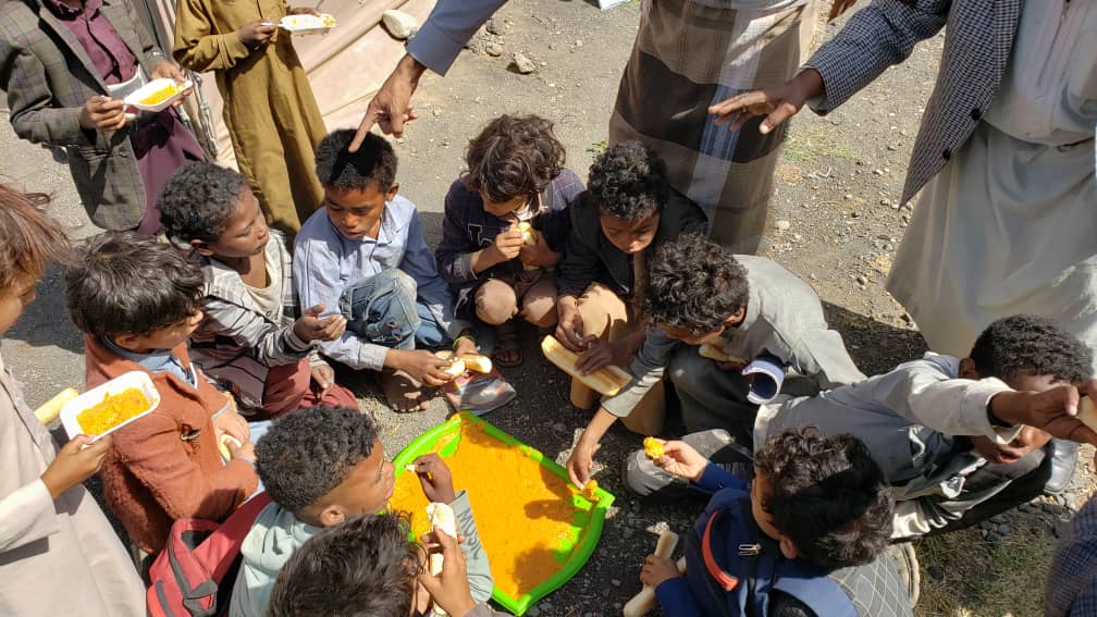 Auf dem Bild ist eine Gruppe Kinder zu sehen, die in einem Kreis sitzen und zusammen Brot essen. In der Mitte steht ein Tablett mit einer orangenen Paste.