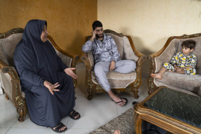 Om Shobi, eine Frau aus dem Gazastreifen, sitzt auf einem Sessel und erzählt etwas. Ein Mann und ein Junge hören ihr zu.