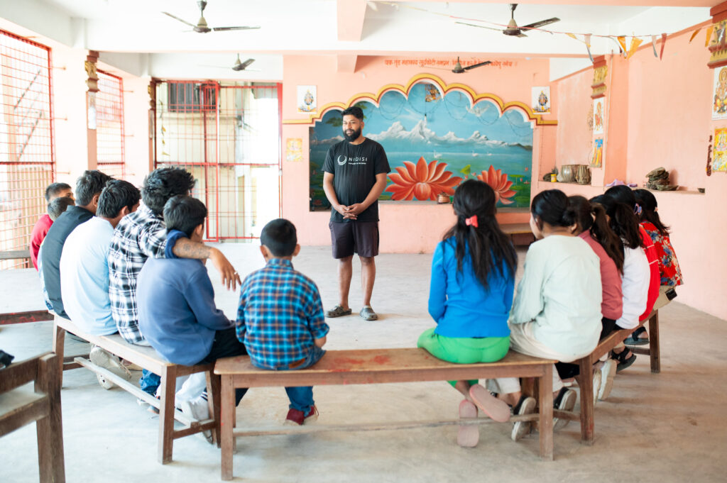 Ein Mitarbeiter von Nidisi in Nepal steht in einem Klassenzimmer vor mehreren Schülern und Schülerinnen unterschiedlichen Alters, die auf Bänken sitzen.