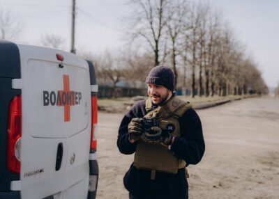Ein ehrenamtlicher Helfer in Schutzweste steht in der Ukraine neben einem Transporter auf dem in kyrillischen Buchstaben "Volunteer" steht.