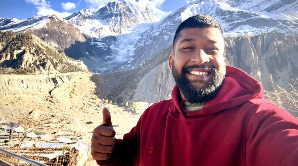 Adhitya, ein junger nepalesischer Mann, steht lachend auf einem Berg in Nepal. Er trägt einen roten Kapuzenpullover und zeigt einen Daumen nach oben. Im Hintergrund sieht man ein Gebirge.