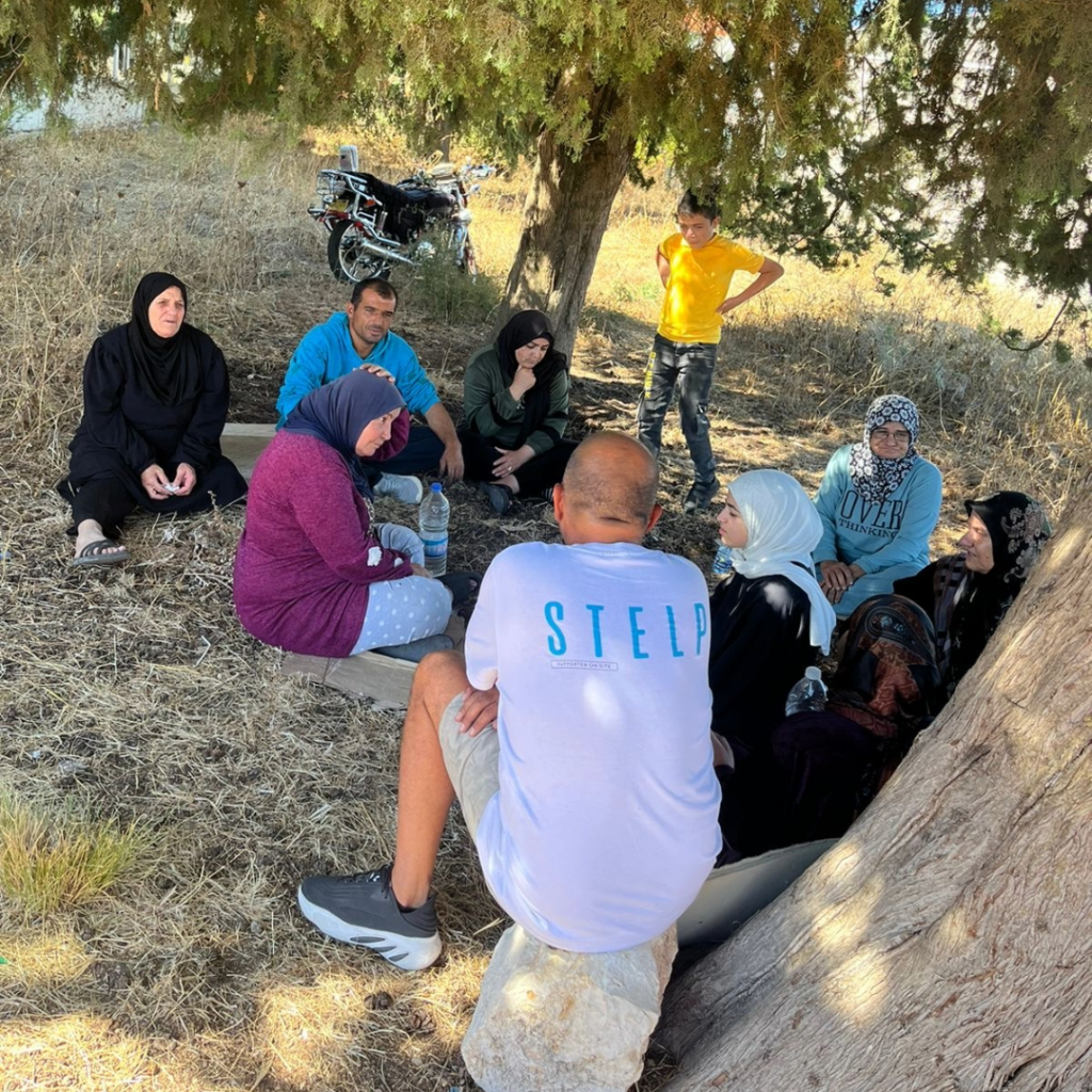 Ein humanitärer Helfer sitzt im Libanon mit binnengeflüchteten Zivilist:innen unter Olivenbäumen zusammen auf dem Boden.